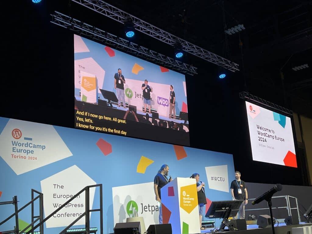 Photo of a stage, three people with microphones in front of a WordCamp Europe branded background