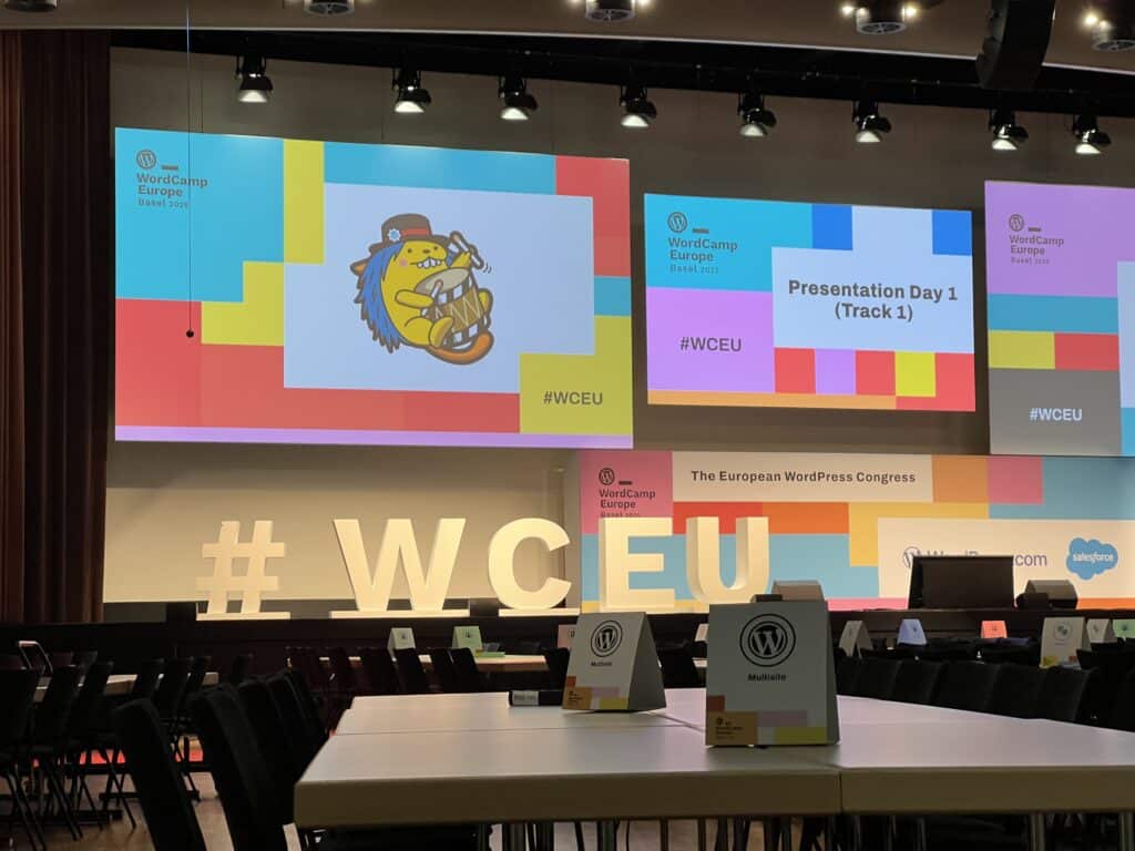 A big stage with multiple projector screens all covered in WordCamp Europe branding. Big letter "#WCEU" proper up on stage. 
In the foreground: tables with signs.