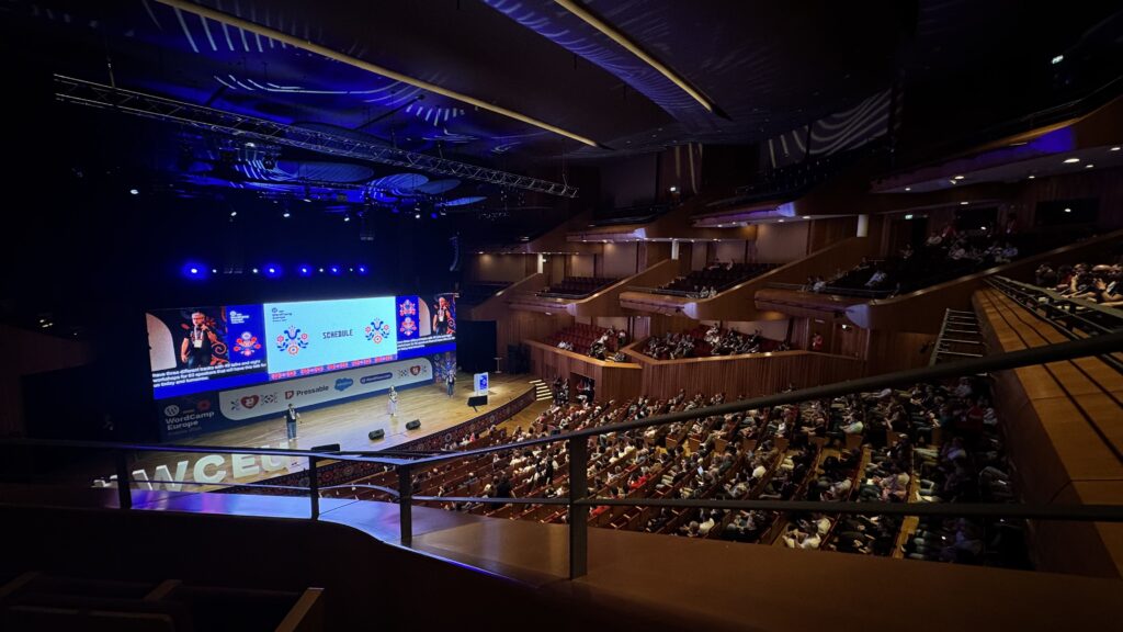 An auditorium full of people. With there people on the stage.