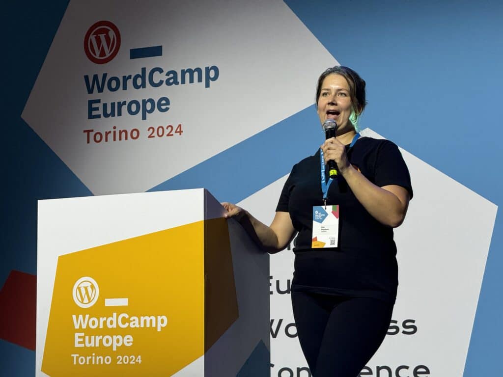 Photo of Liza Bogatyrev on stage with a mic in her hand. The wall behind her shows the WordCamp Europe Logo.